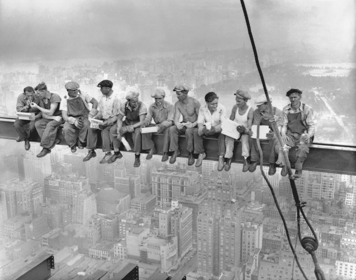 A black-and-white historical photograph showing eleven construction workers sitting on a steel beam high above a city skyline, eating lunch and holding newspapers. The image is the famous 1932 photograph of workers on the Empire State Building.