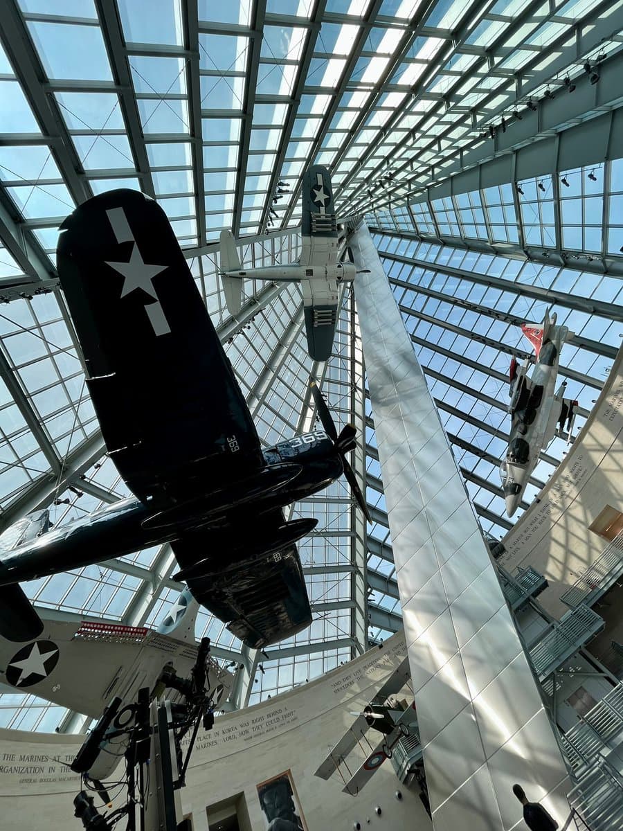 A low-angle photograph taken inside a large aviation museum with a glass ceiling, showing several vintage military aircraft suspended from the ceiling. The closest aircraft is a dark-colored propeller plane with a white star insignia on its wing, while other planes are visible in the background.