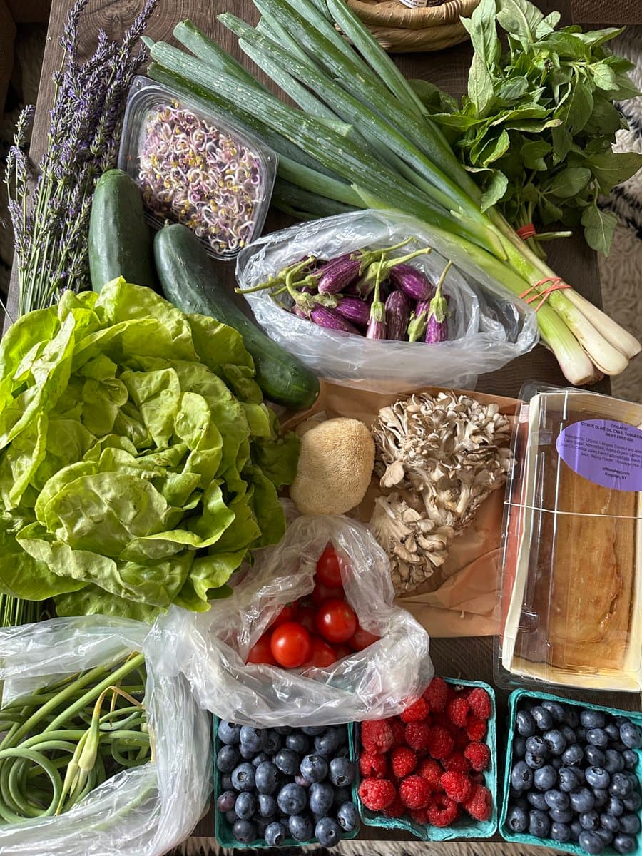 A top-down photograph of a variety of fresh produce, including lettuce, zucchini, leeks, and berries, arranged on a wooden table. The assortment also includes packaged mushrooms, tomatoes, and a container of sprouts, suggesting a recent harvest or market purchase.