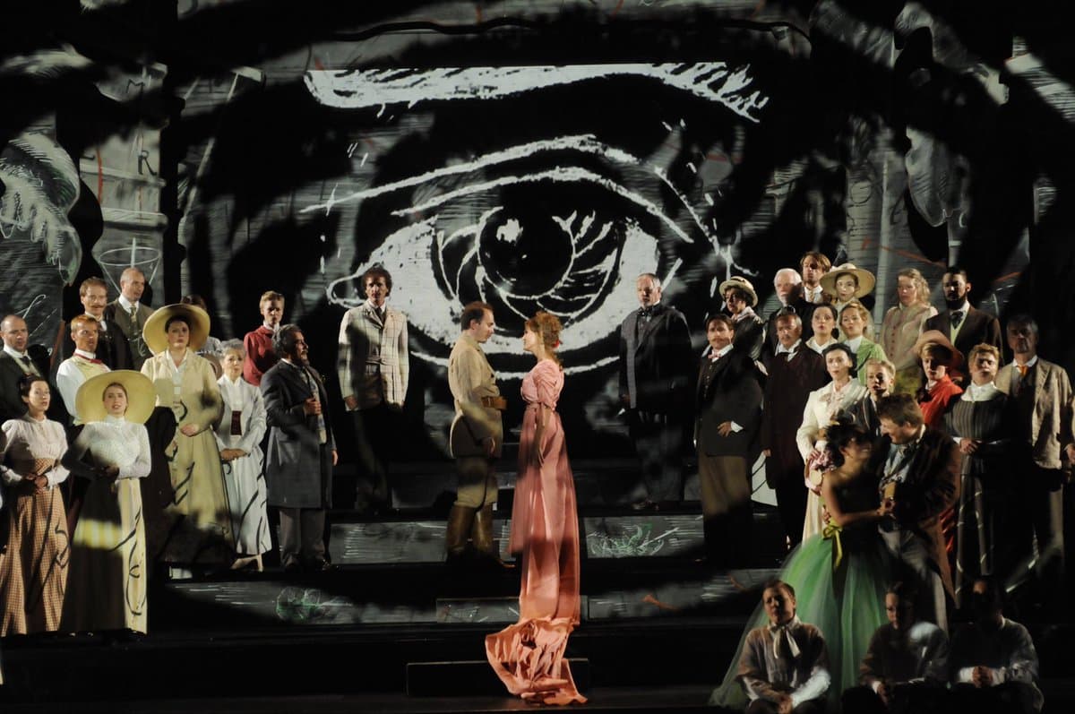 A wide shot of an opera performance featuring a large cast of actors in period costumes on a stage. A woman in a long pink dress stands at the center facing a man, while the rest of the cast is arranged on tiered platforms behind them against a dark backdrop featuring a large, stylized white eye.