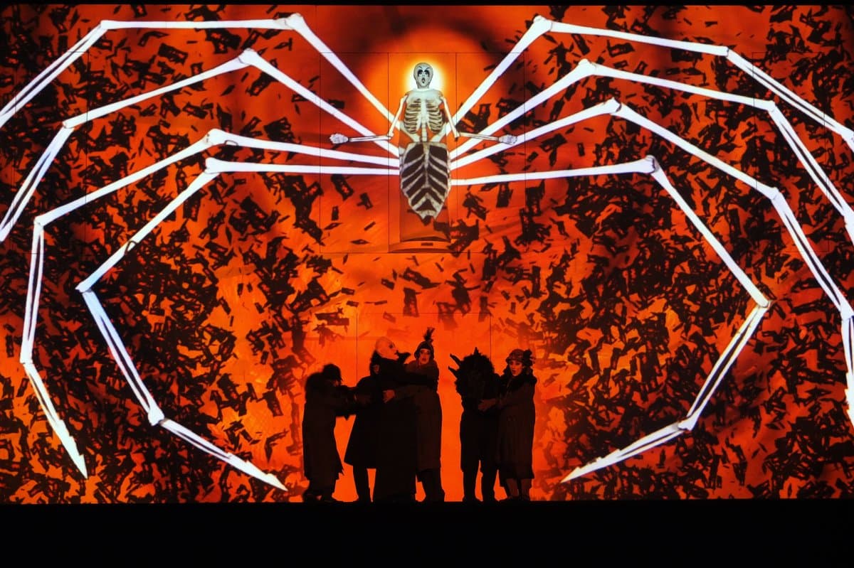 A theatrical stage production featuring a large, projected image of a skeleton with spider-like legs against a fiery orange background, with several performers in dark costumes standing on the stage below.
