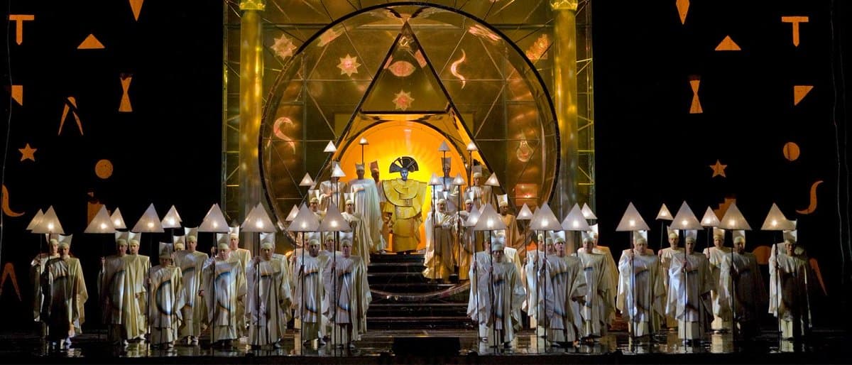 A wide shot of an opera stage featuring a large choir in white robes holding cone-shaped lamps, with a central figure in gold robes standing on a raised platform beneath a large, illuminated geometric structure.