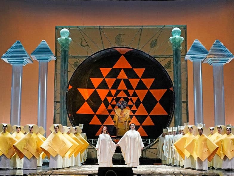 A wide shot of a theatrical stage production featuring a large, circular backdrop with a black and orange geometric triangle pattern. In the foreground, two performers in white robes stand center stage, flanked by a chorus of singers wearing yellow and white costumes, with stylized blue and green pillars in the background.
