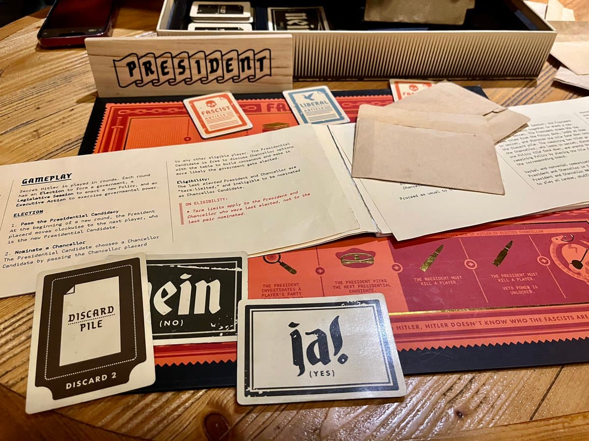 A photograph of a Secret Hitler board game in progress, showing the game board, rulebook, and various game components on a wooden table. The setup includes a wooden "PRESIDENT" sign, role cards for Fascist and Liberal, and voting cards labeled "ja!" (Yes) and "Nein" (No).