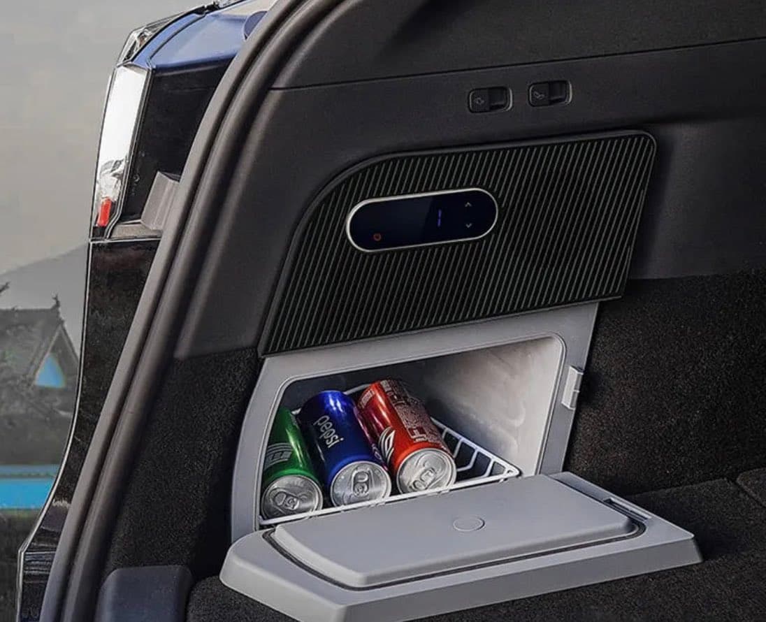 A close-up view of a car trunk featuring a built-in mini-fridge with its door open, revealing three soda cans inside. Above the fridge is a black panel with a digital temperature display and control buttons.