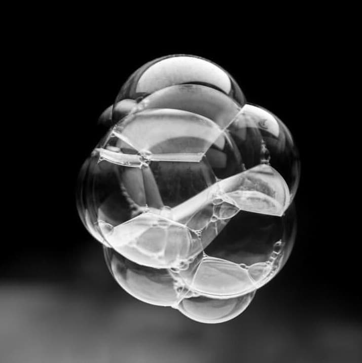 A black-and-white photograph of a cluster of transparent, spherical bubbles or droplets against a dark background. The image highlights the geometric shapes and reflections within the individual spheres.