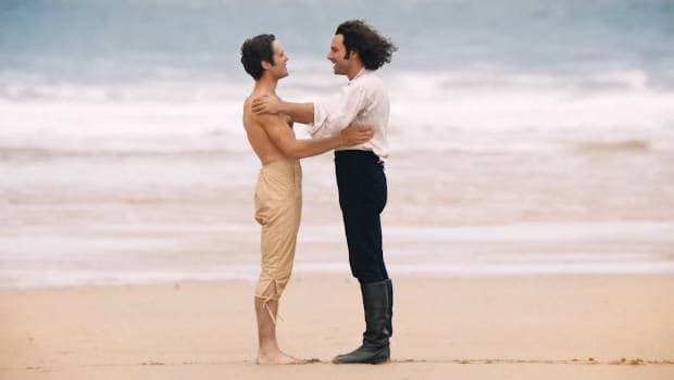 Two people stand on a sandy beach facing each other, with one person shirtless and the other wearing a white shirt and dark pants, set against a backdrop of ocean waves.