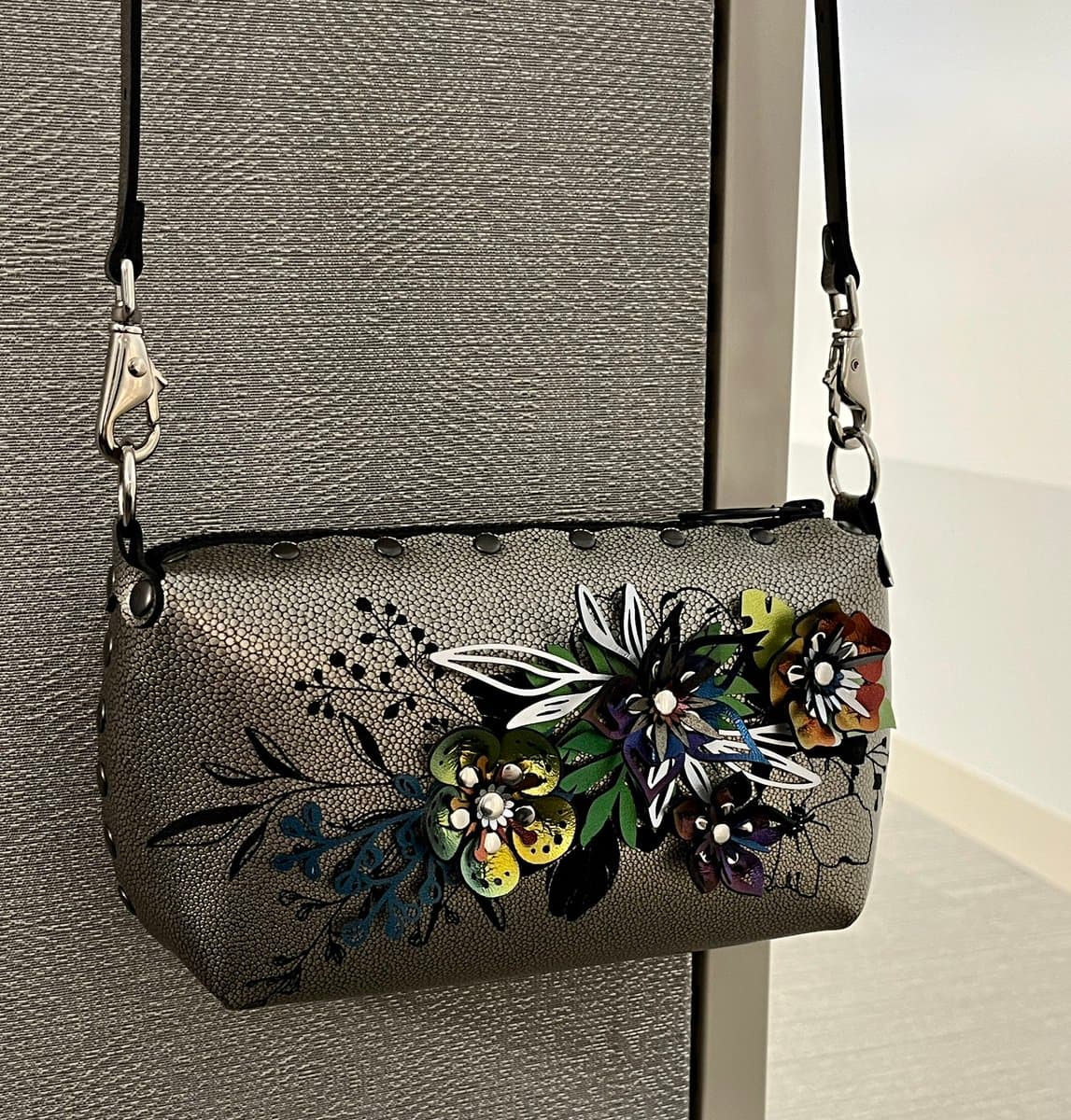 A close-up photograph of a textured, grey handbag with a black strap hanging against a grey wall. The bag features a prominent, colorful floral design with metallic and beaded details on the front.