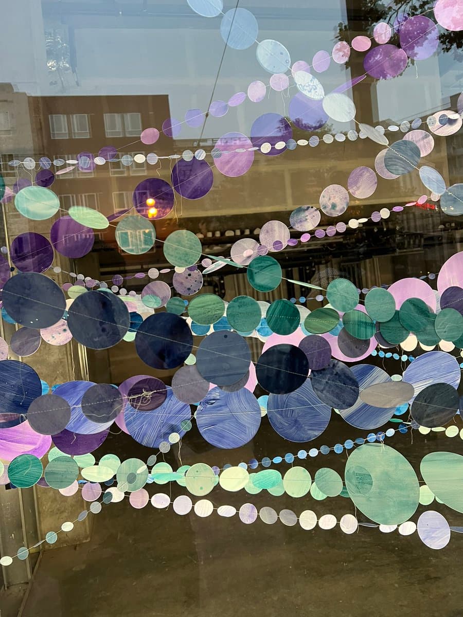 A photograph of a window display featuring numerous circular paper cutouts in shades of blue, purple, and green strung on threads. The decorations are arranged in overlapping layers against a glass pane that reflects a building across the street.
