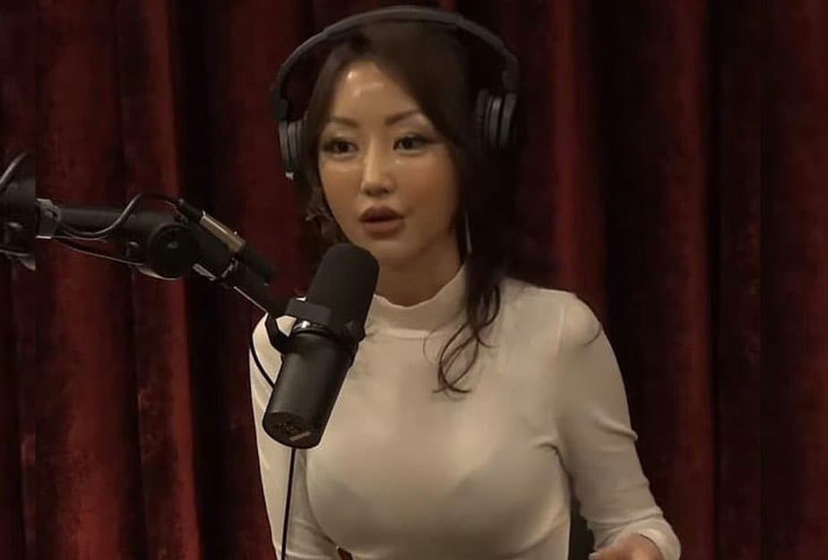 A woman wearing headphones and a white turtleneck sits in front of a professional microphone, appearing to be in a recording or podcasting session. She is positioned against a dark red curtain background.