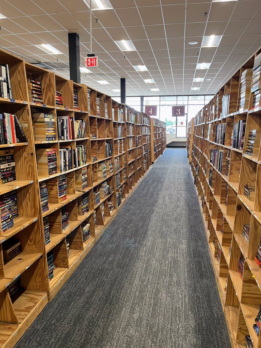 A wide-angle photograph of a long aisle in a used bookstore, flanked by wooden shelves filled with books and media. The aisle is carpeted in grey and leads toward a window at the far end, with numbered signs hanging from the ceiling.