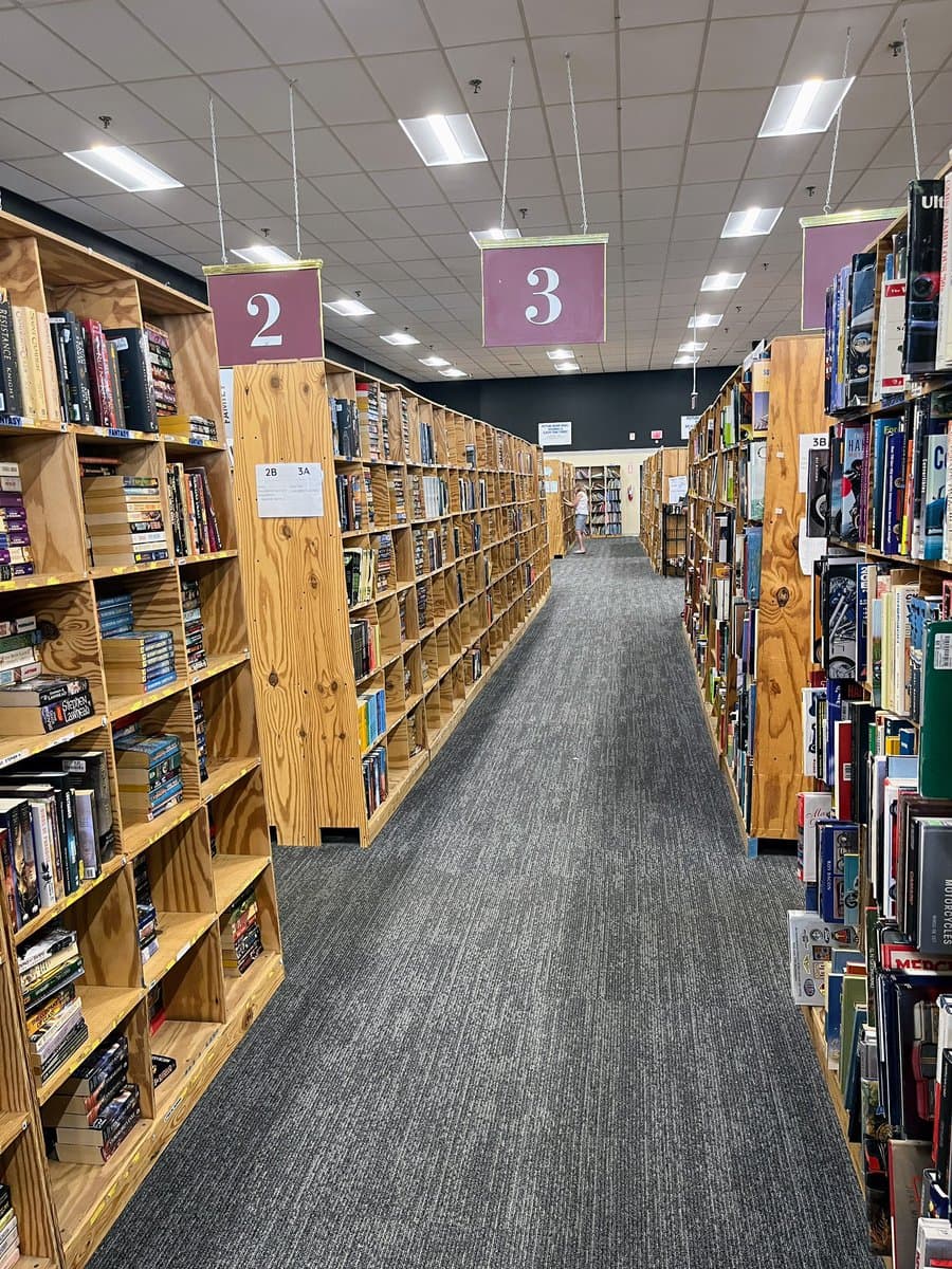 A wide-angle photograph of a long aisle in a used bookstore, featuring wooden bookshelves packed with books on both sides and purple hanging signs numbered 2 and 3 above the shelves.