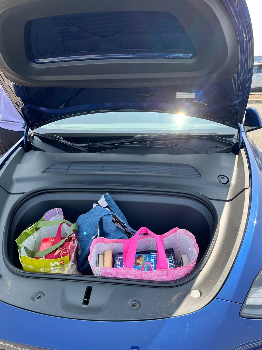 A photo of the open front trunk (frunk) of a blue car, filled with several reusable shopping bags containing books. The scene is brightly lit by sunlight reflecting off the car's windshield.