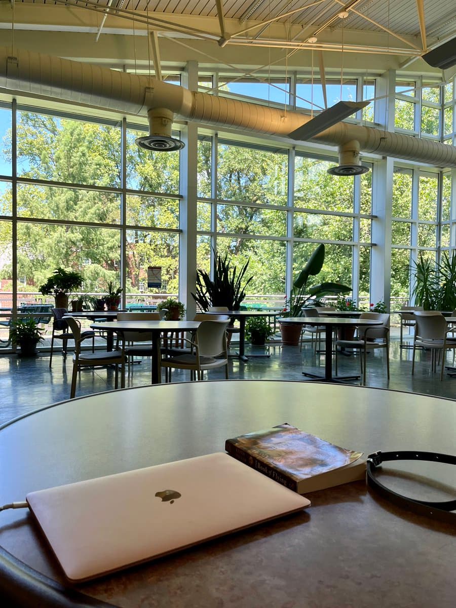 A first-person view of a laptop, a book, and a watch on a table inside a bright, modern community center with large windows and potted plants.
