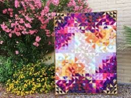A colorful geometric quilt featuring a pinwheel pattern in shades of purple, orange, and white, leaning against a wooden fence next to a bush with pink flowers and yellow ground cover.