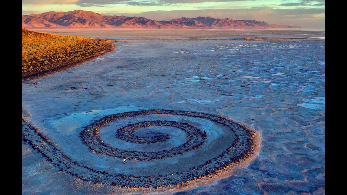 A landscape photograph of the Spiral Jetty art installation, a spiral-shaped structure made of rocks extending into a shallow, cracked lake bed. In the background, a mountain range is visible under a soft, pastel-colored sky at either sunrise or sunset.