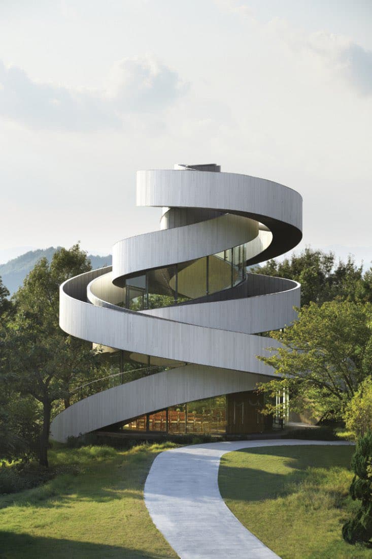 A photograph of the Ribbon Chapel, a modern building with a distinctive white, spiraling ribbon-like structure that wraps around a central glass-walled core. The building is situated on a grassy hillside surrounded by trees, with a winding concrete path leading to its entrance.