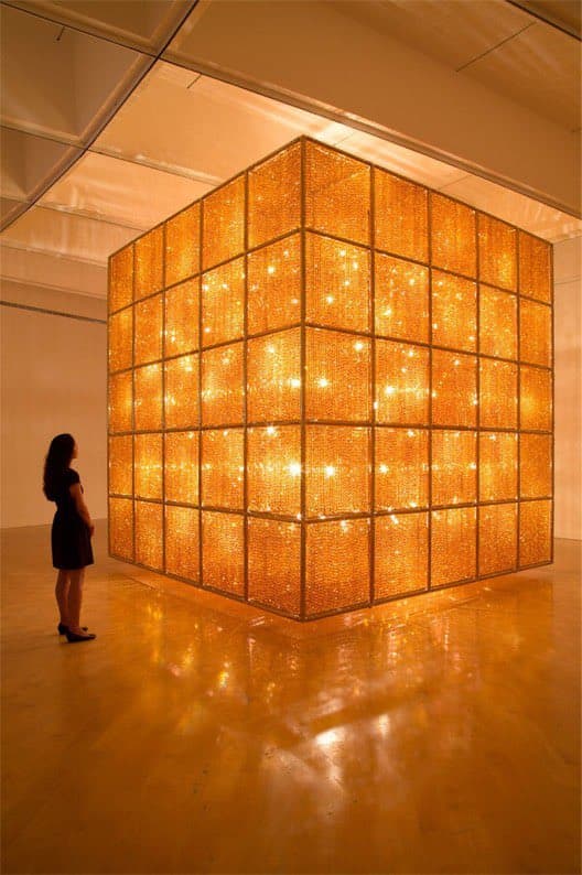 A large, illuminated cube art installation composed of a grid of glowing, textured panels stands in a gallery space, with a person standing to the left for scale.
