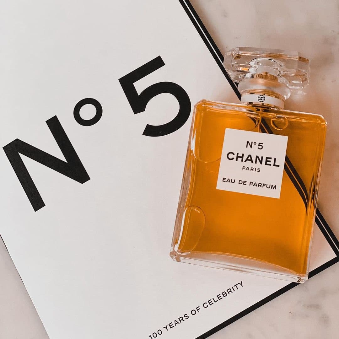 A bottle of Chanel No. 5 Eau de Parfum sits on a white surface next to a book or card featuring the text "N°5" and "100 YEARS OF CELEBRITY".