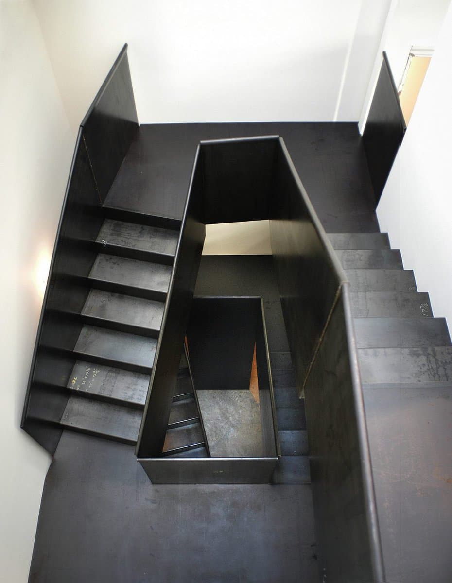 A top-down view of a modern, industrial-style staircase with dark metal steps and railings set against white walls. The structure features a central void and angular design elements.