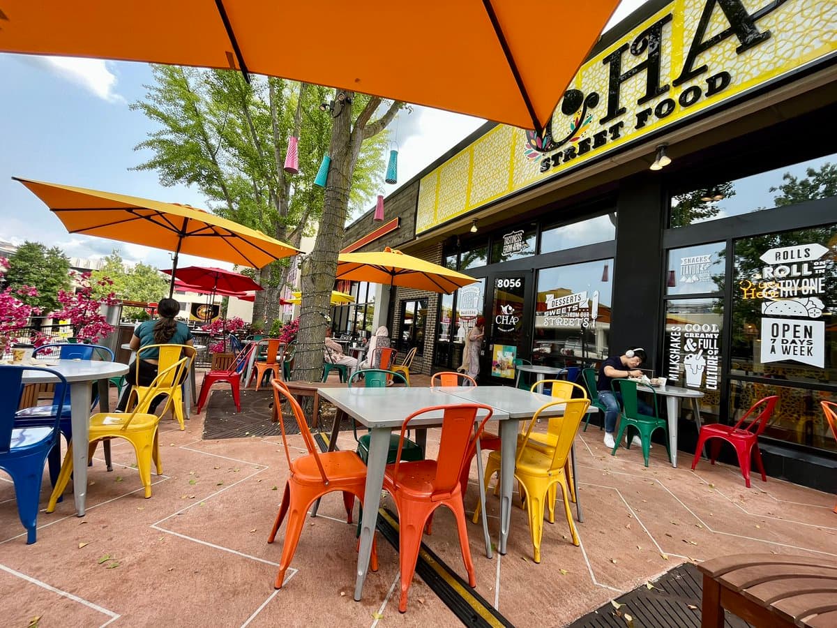 An outdoor dining area at a restaurant called Cha Street Food, featuring colorful metal chairs and tables under orange umbrellas. The storefront has large windows with various food-related signs and a yellow sign above the entrance.
