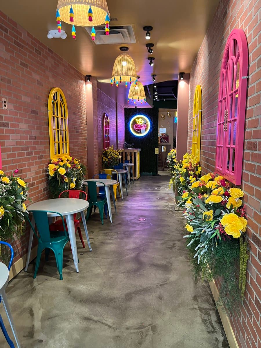 A brightly lit, narrow restaurant interior with exposed brick walls, colorful wicker pendant lights, and decorative faux windows in yellow and pink. The space is decorated with large arrangements of yellow flowers and features small round tables with colorful metal chairs. A neon sign reading "CHA" is visible at the far end of the corridor.