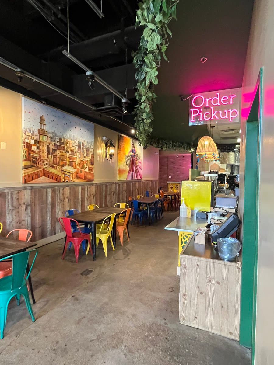 A photo of the interior of a street food restaurant featuring colorful metal chairs, wooden tables, and wall murals of a cityscape and a person. A pink neon sign reading "Order Pickup" is visible above a service counter on the right.