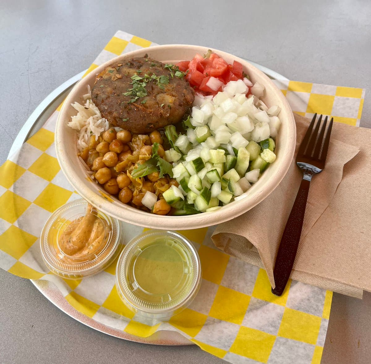 A top-down view of a takeout meal served in a paper bowl on a yellow-and-white checkered napkin, featuring a falafel patty, rice, chickpeas, diced tomatoes, onions, and cucumbers, accompanied by two small containers of sauce and a plastic fork.