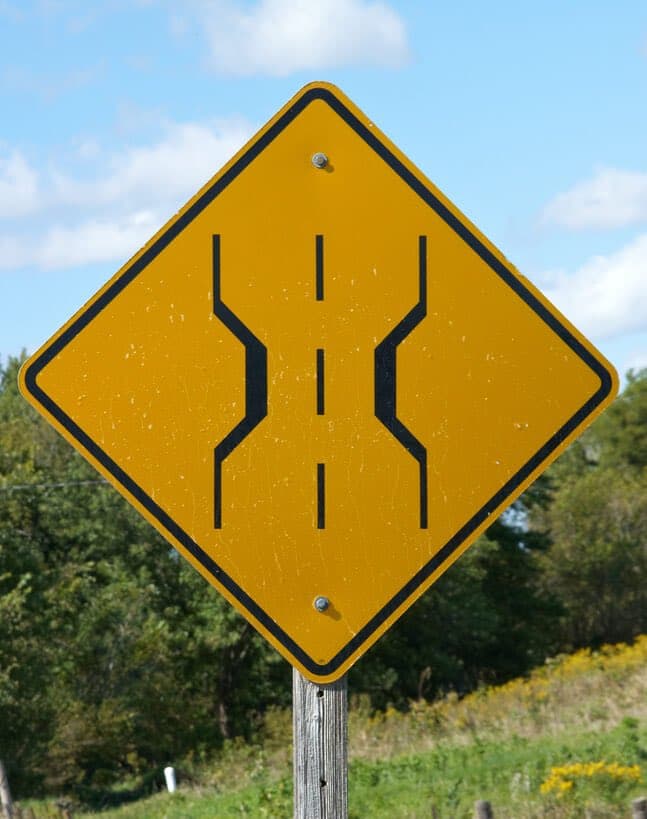 A yellow diamond-shaped road sign on a wooden post, depicting a narrow bridge symbol with a road narrowing in the middle, set against a backdrop of green trees and a blue sky with clouds.