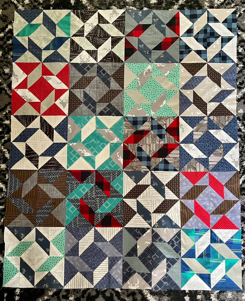 A top-down view of a finished quilt top laid flat on a dark, textured surface. The quilt is composed of a grid of square blocks, each featuring a geometric pattern made from triangles in various fabrics, including plaid, deer prints, and solid colors in red, blue, green, and brown.