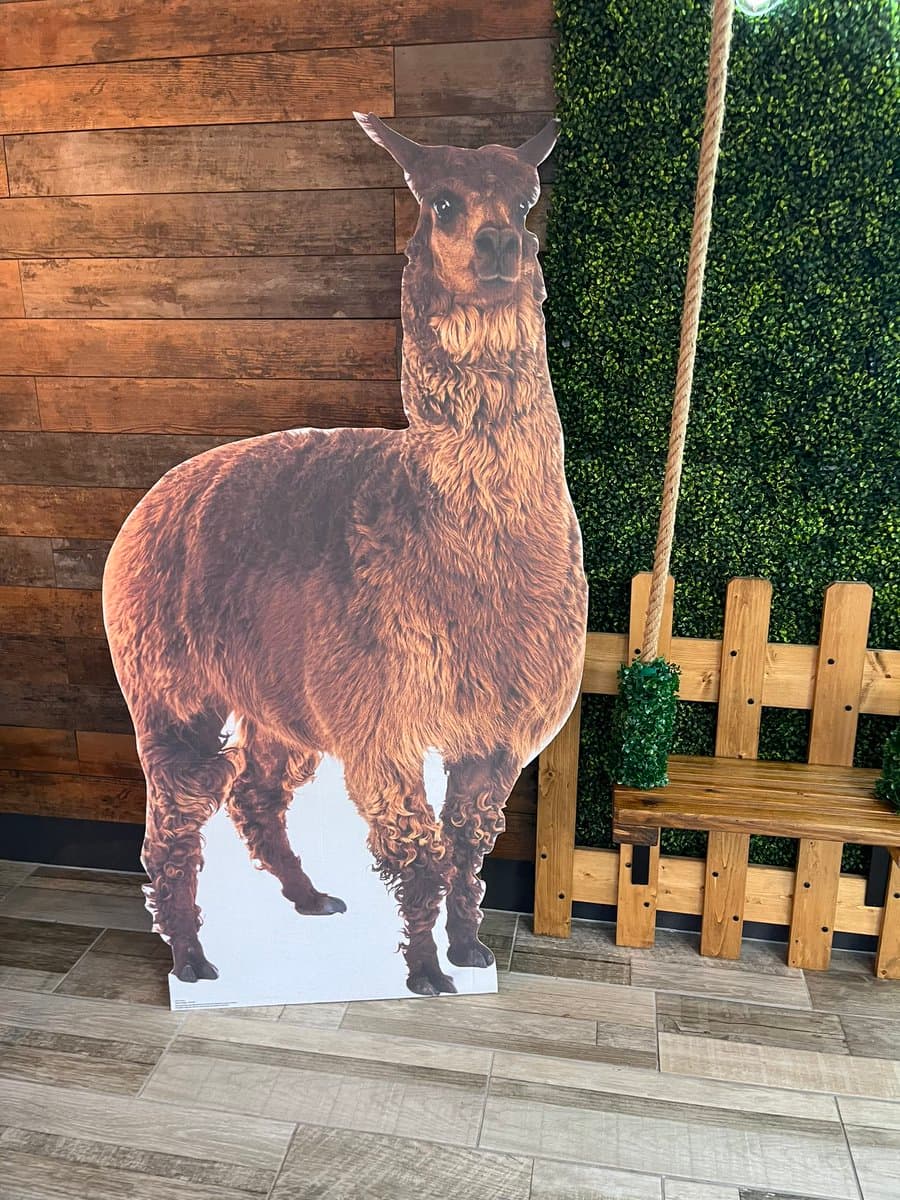 A life-sized cardboard cutout of a brown llama stands on a tiled floor in front of a wooden wall and a green hedge, next to a wooden swing.