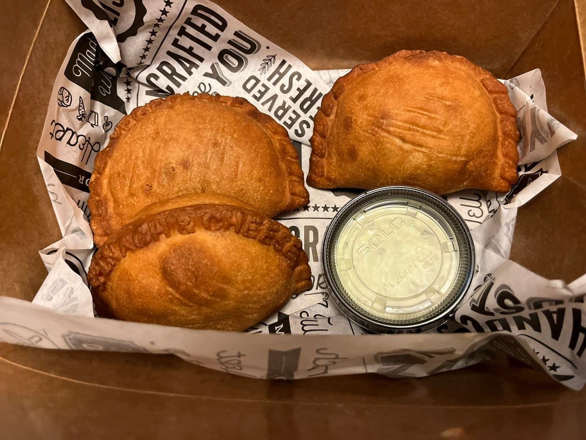 A top-down view of a cardboard takeout box containing three golden-brown, baked empanadas and a small plastic container of white dipping sauce, resting on branded paper.