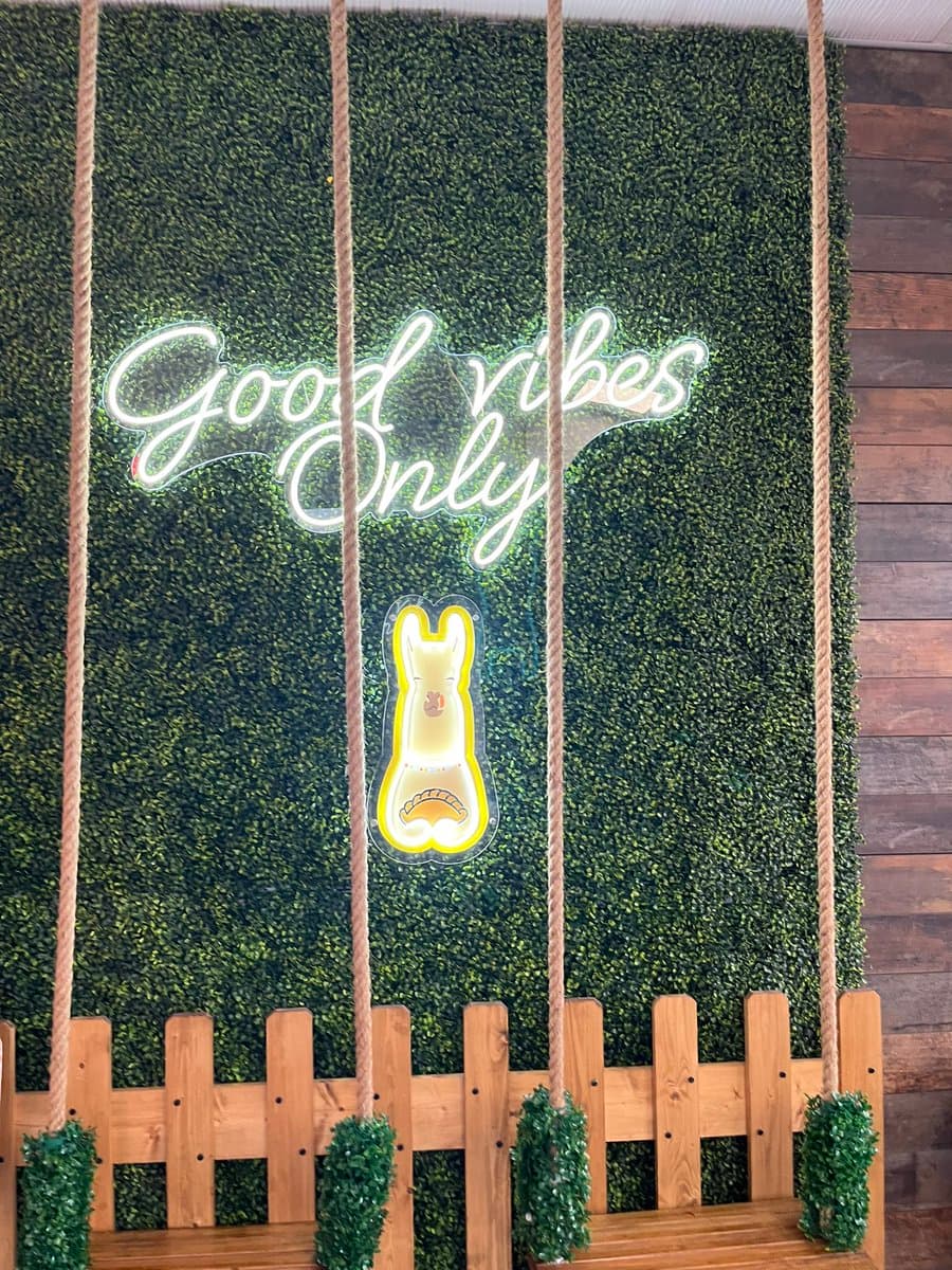 A wooden swing set with greenery-wrapped ropes hangs in front of a living green wall. A white neon sign on the wall reads "Good vibes Only" above a yellow neon outline of a dog.