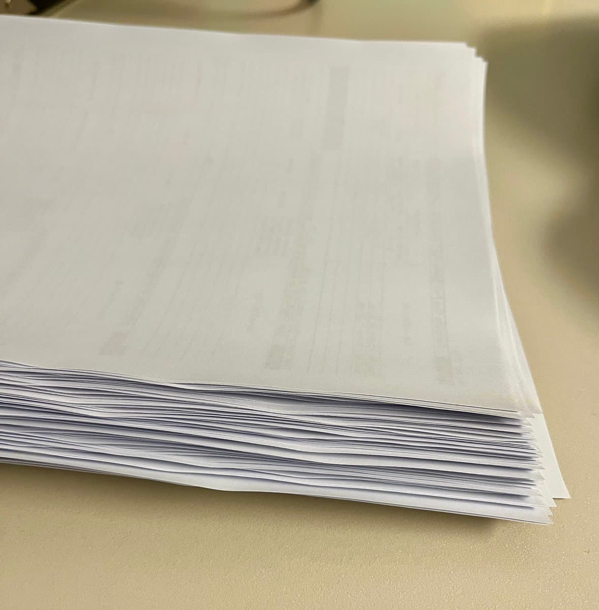 A thick stack of printed white paper documents rests on a light-colored surface. The pages are slightly fanned out, revealing their edges, while the top sheet shows faint, illegible text patterns typical of a form.