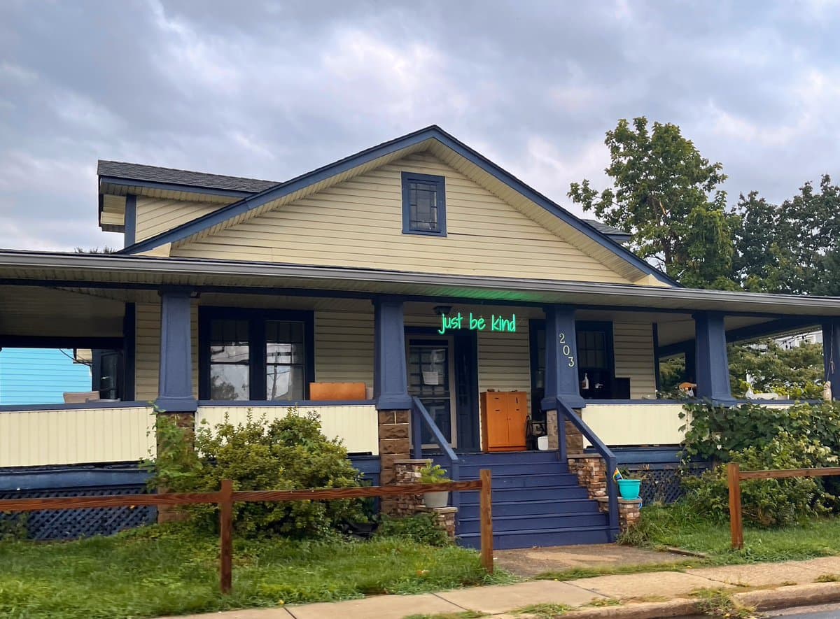 A photograph of a single-story house with light yellow siding and dark blue trim, featuring a front porch with a green neon sign that reads 'Just be kind' and the house number '203' on a pillar. The house is surrounded by greenery and a wooden fence under a cloudy sky.