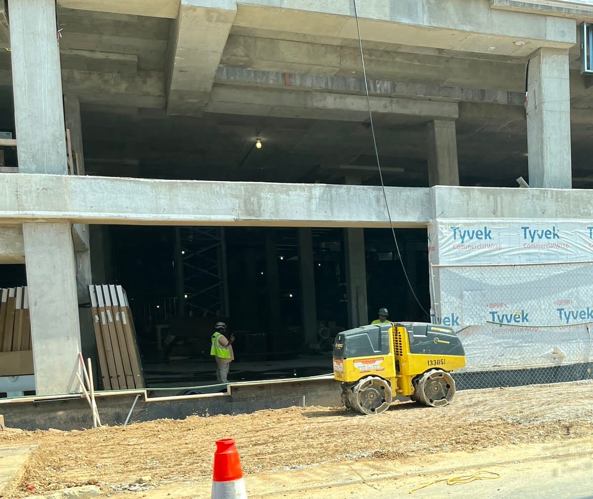 A construction site featuring a small yellow and black Bobcat skid-steer loader parked on a dirt surface in front of a concrete building under construction. Two workers in high-visibility vests are visible near the building's open interior, which is partially covered with white Tyvek CommercialWrap.