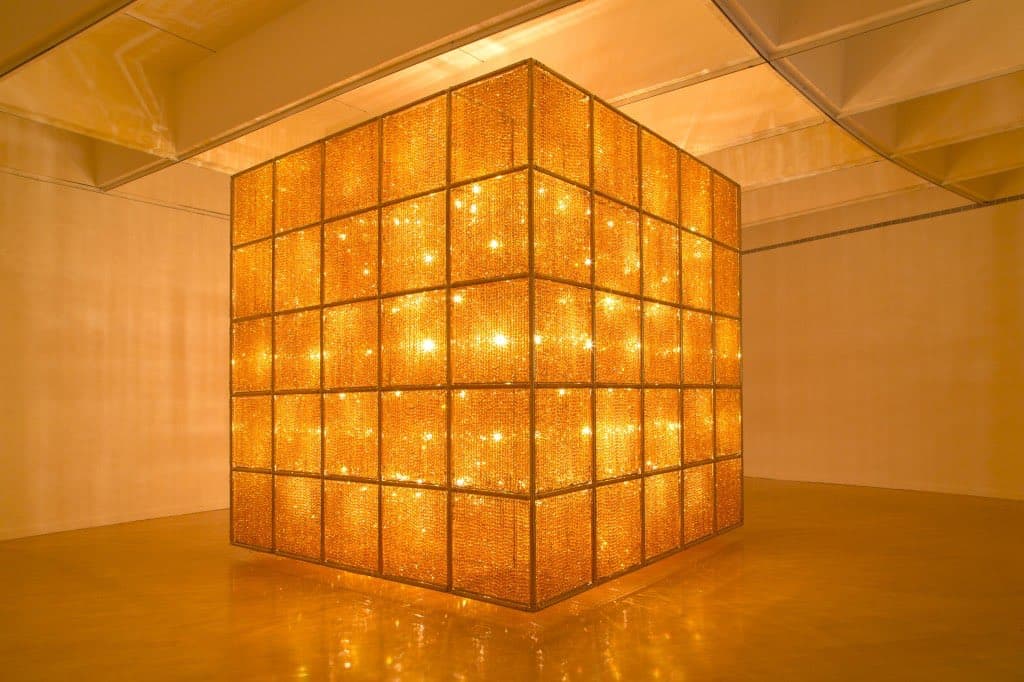 A large, illuminated cube sculpture made of a grid of glowing, textured panels stands in the center of a minimalist gallery room with warm lighting. The structure is positioned on a polished floor that reflects its golden light.