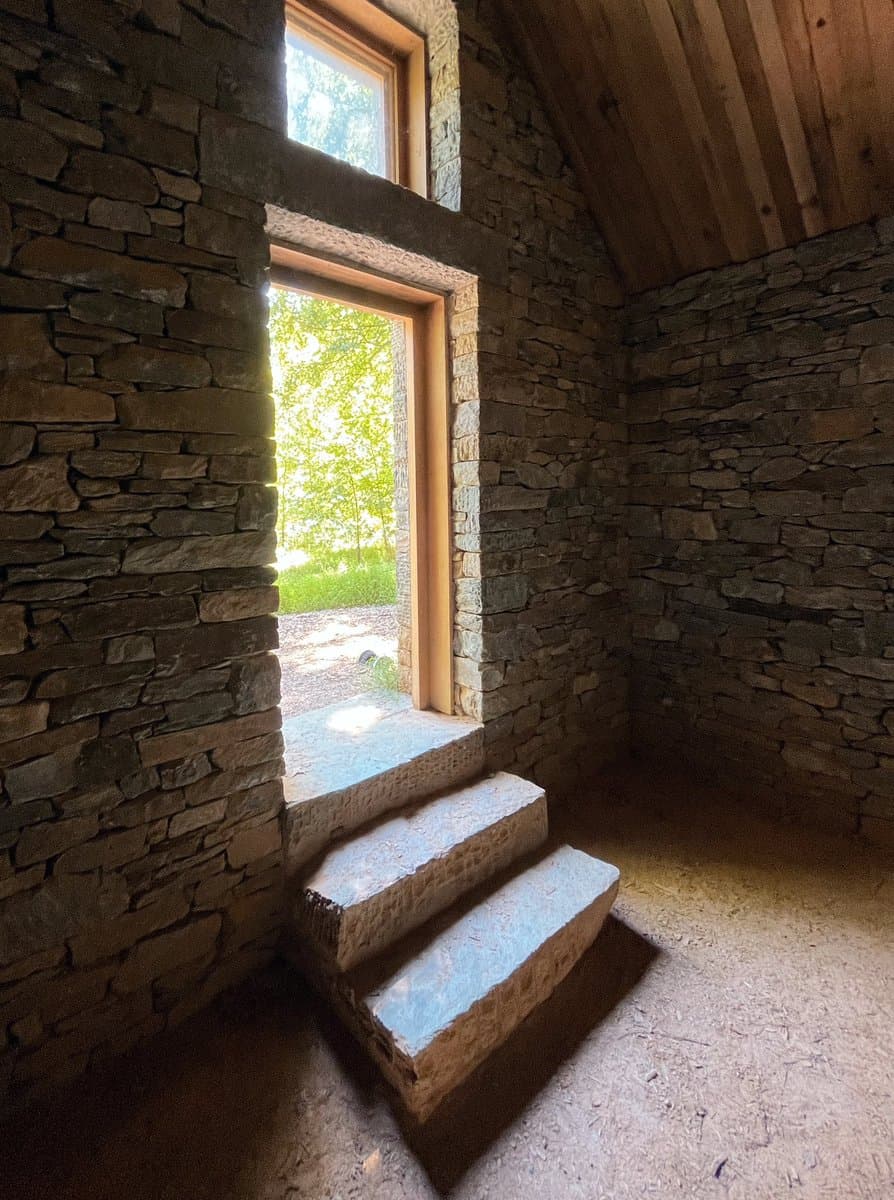 An interior view of a rustic room with walls made of stacked, irregular stones and a wooden plank ceiling. A set of three rough-hewn stone steps leads up to a window opening that looks out onto a bright, green outdoor area.