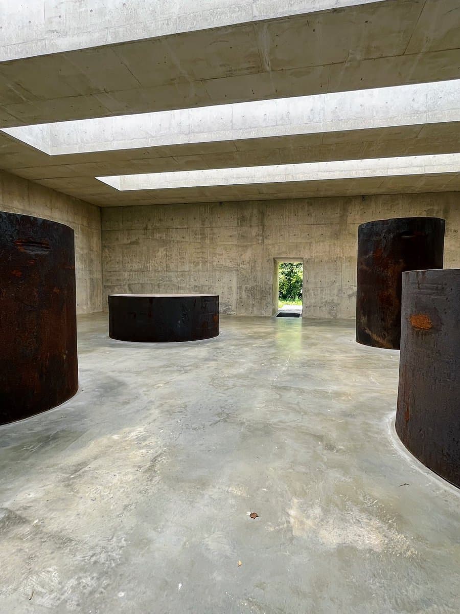 A minimalist concrete interior space featuring a polished floor, exposed concrete walls, and a ceiling with rectangular skylights. Several large, rusted cylindrical metal structures are arranged throughout the room, with a small doorway in the back wall revealing greenery outside.