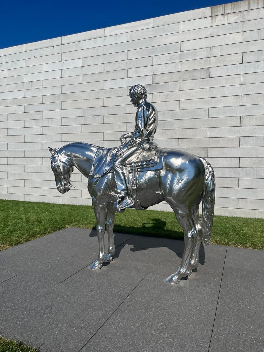 A shiny, metallic silver sculpture of a person riding a horse, positioned on a paved surface in front of a white stone wall and green grass under a clear blue sky.