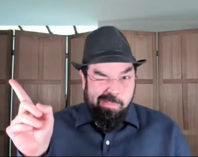 A man with a beard, wearing a dark hat and a blue button-down shirt, is shown in a close-up shot. He is winking and pointing his index finger upward while making a facial expression, set against a background of wooden closet doors.