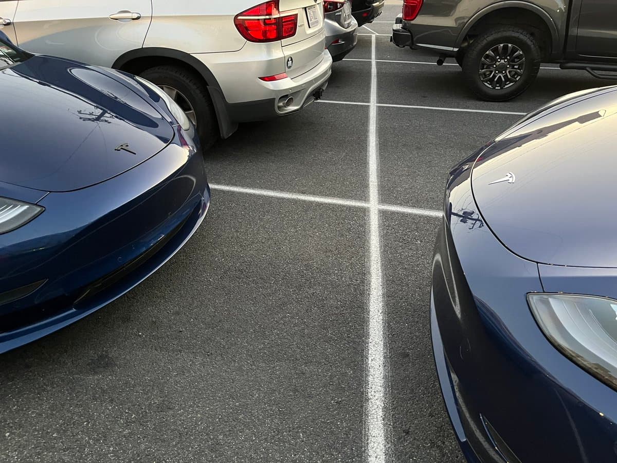 A photo taken in a parking lot showing the front hoods of two blue Tesla vehicles, highlighting the difference between a black logo on the left and a chrome logo on the right.
