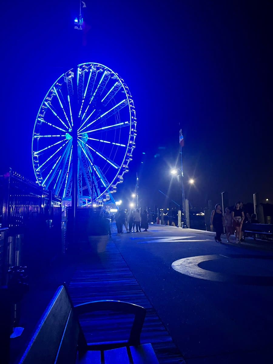 A nighttime photograph of a pier at National Harbor, featuring a large Ferris wheel illuminated with bright blue lights. Several people are walking along the pier, which is lit by streetlamps, with a bench visible in the foreground.
