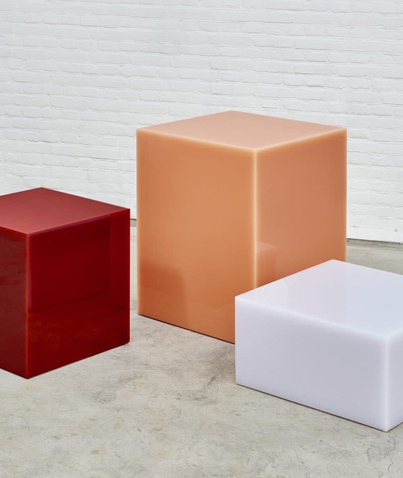 A photograph of three translucent, cube-shaped stools in red, peach, and white, arranged on a concrete floor against a white brick wall.