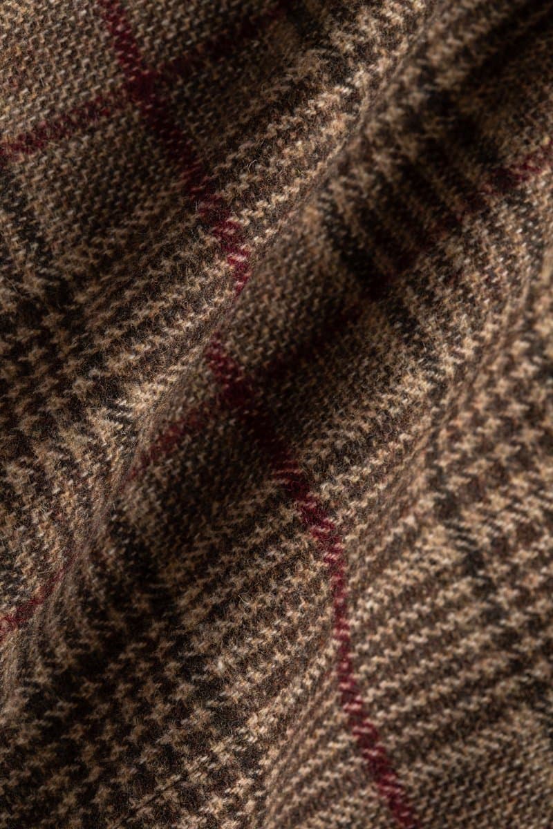 A close-up photograph of a textured, brown plaid fabric with thin red and white lines. The material appears to be a woven wool or tweed, shown in soft folds.