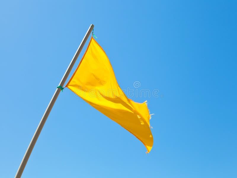 A yellow flag attached to a pole is waving in the wind against a clear blue sky. The flag appears to be made of a lightweight fabric and is secured to the pole with green ties.