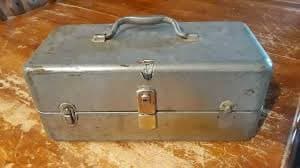 A vintage, rectangular metal toolbox with a handle and latches sits on a wooden surface. The toolbox appears to be made of galvanized steel and shows signs of wear and age.