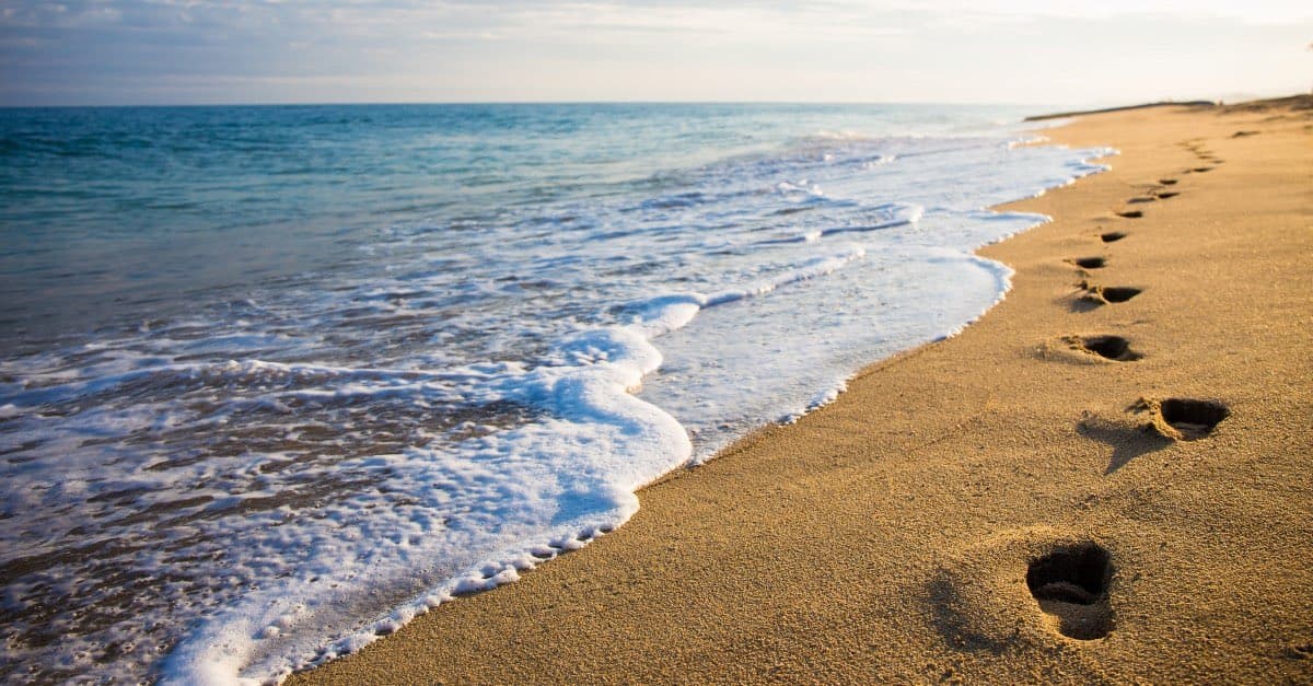 A photograph of a sandy beach with gentle waves lapping at the shore, featuring a trail of footprints leading into the distance along the water's edge.
