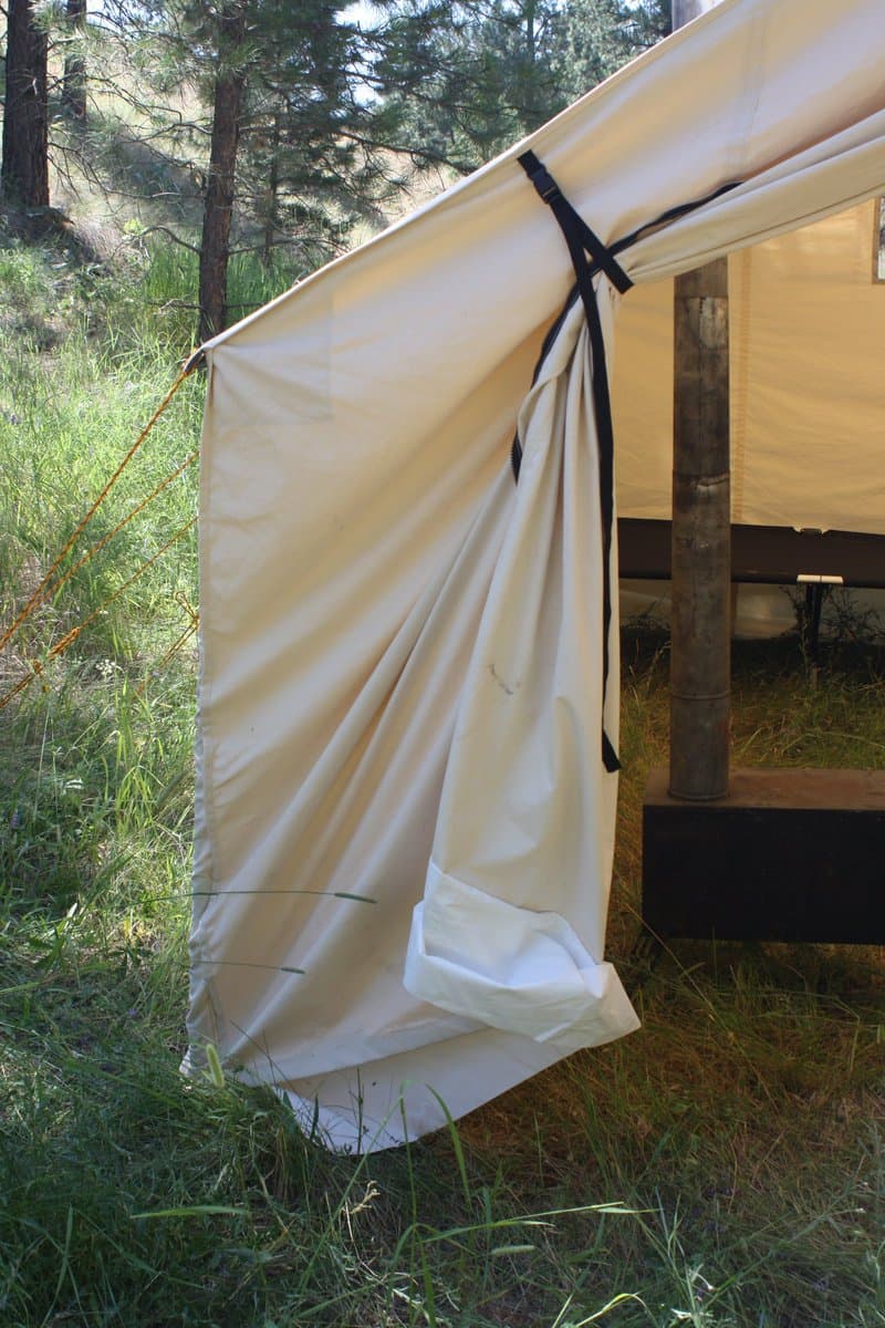 A canvas tent is set up in a grassy, wooded area, with its side flap tied back to reveal the interior. The tent is supported by a wooden pole and secured with guy lines.