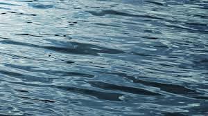 A close-up photograph of the surface of a body of water, showing gentle ripples and waves in shades of blue and gray. The image captures the texture and movement of the water without any visible land or objects.